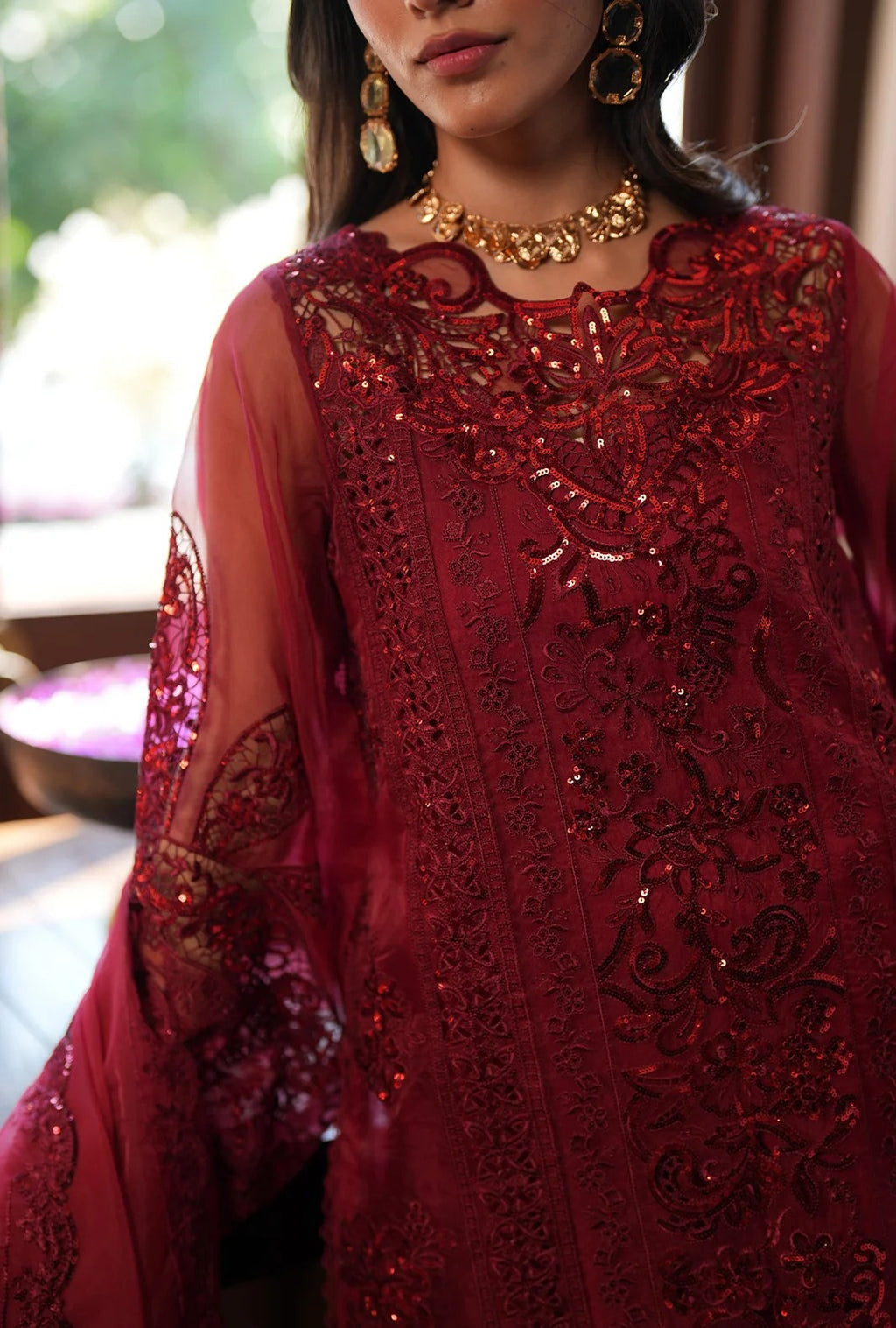 A close-up of a woman wearing a rich red embroidered traditional outfit with sheer sleeves, intricate sequin and lace detailing, and gold jewelry.