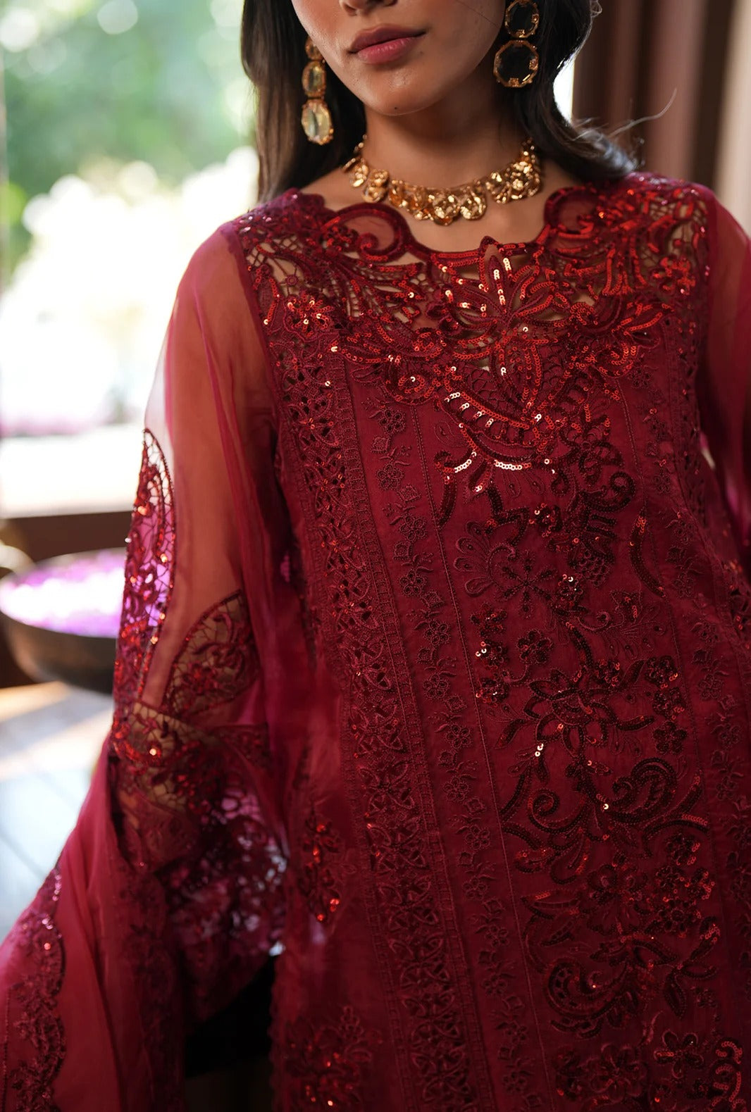 A close-up of a woman wearing a rich red embroidered traditional outfit with sheer sleeves, intricate sequin and lace detailing, and gold jewelry.