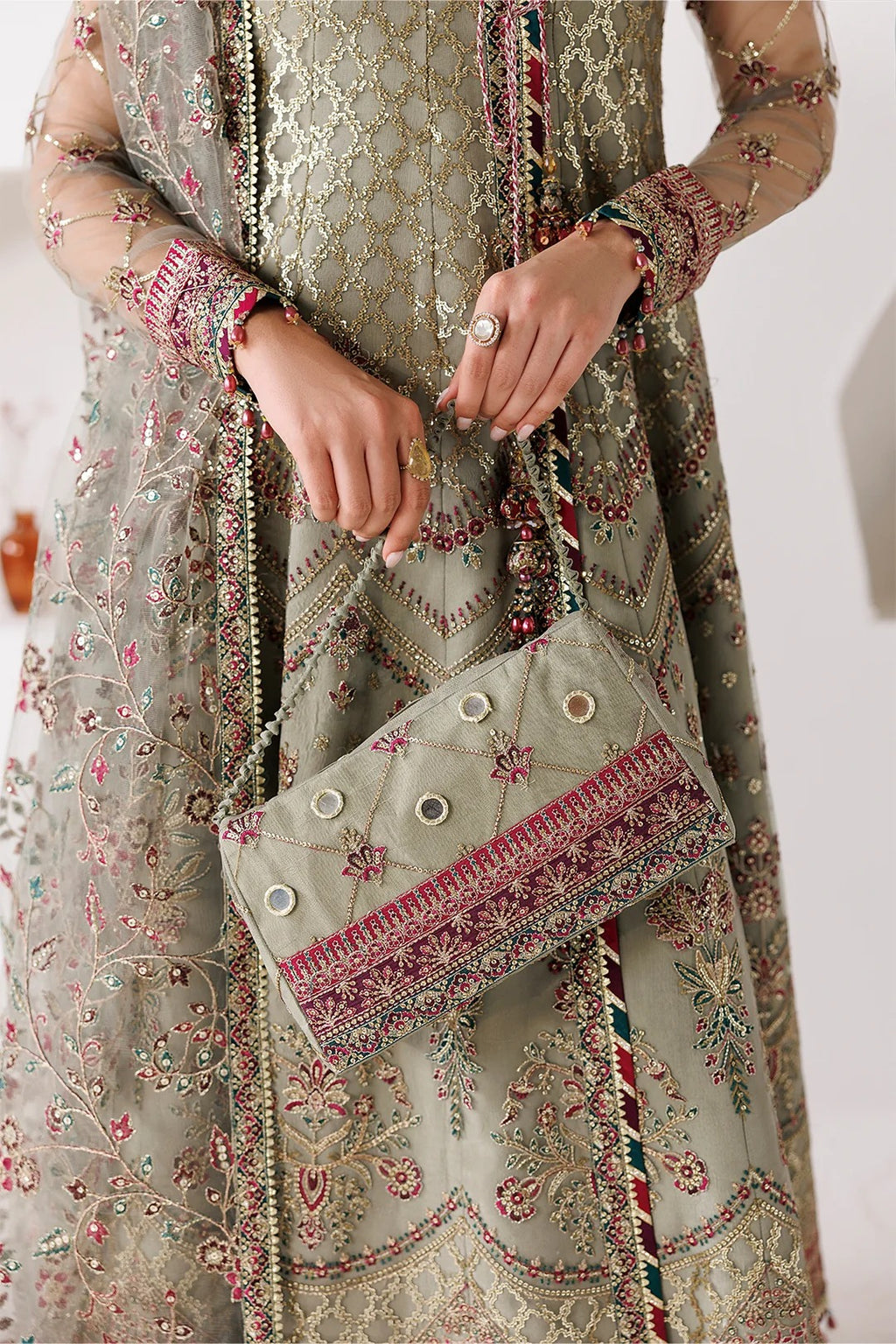 A close-up of a person wearing an ornate olive green embroidered traditional outfit with pink and gold detailing, holding a matching embroidered clutch bag.