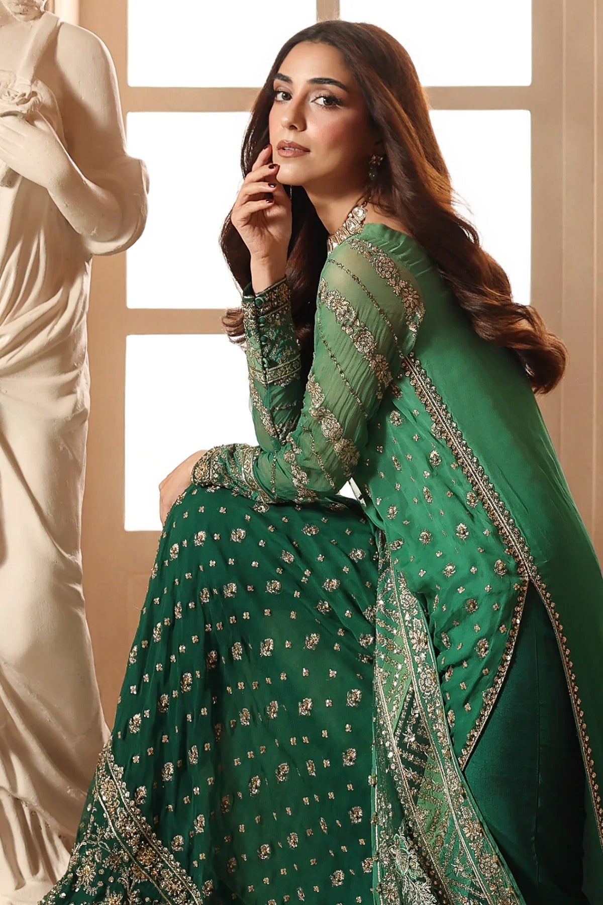 A woman models a green traditional outfit with intricate gold embroidery and a matching draped scarf in a softly lit indoor setting.