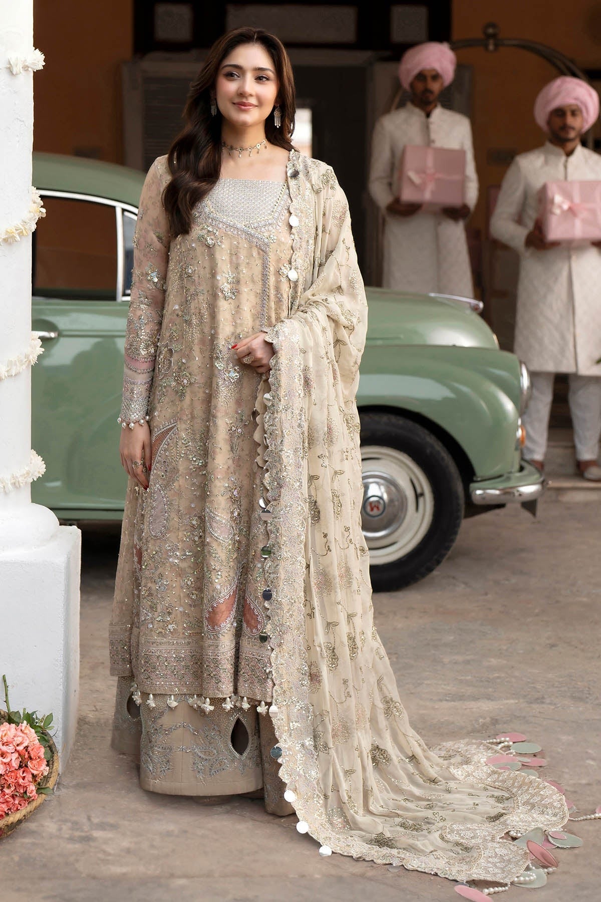 A woman models a traditional beige and gold embroidered Indian ethnic suit with a matching dupatta, standing in front of a vintage green car with men in the background.