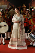A woman in an ornate cream and pink embroidered traditional outfit stands on a red carpet, surrounded by a brass band in red uniforms against a dark illustrated backdrop.
