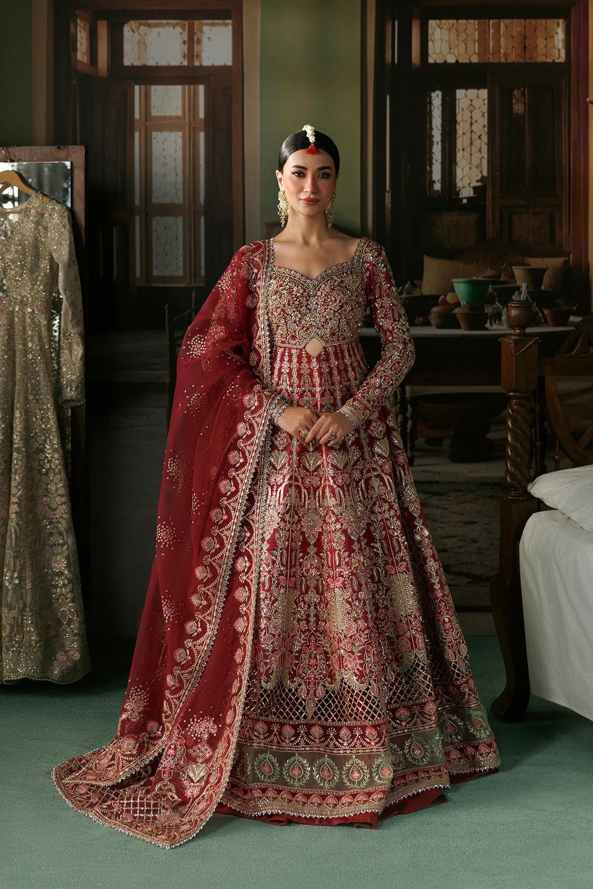 A woman stands indoors wearing an ornate red and gold embroidered traditional outfit with a long draped dupatta. The setting appears to be a furnished room with wooden doors and furniture in the background.
