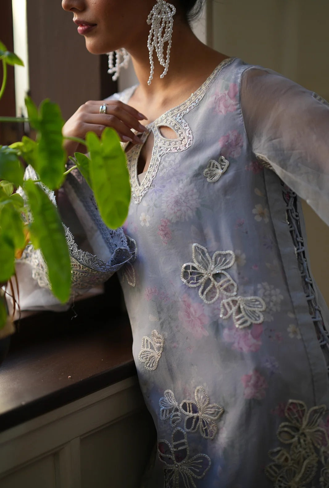 A woman wearing a light blue floral embroidered tunic with sheer sleeves and intricate lace detailing, shown in a softly lit indoor setting with green leaves in the foreground.