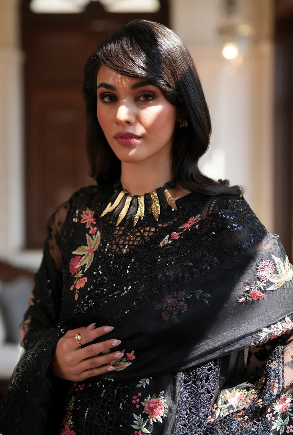 A woman wearing a black embroidered traditional outfit with floral detailing and a matching sheer shawl, styled with a gold collar necklace, in a softly lit indoor setting.