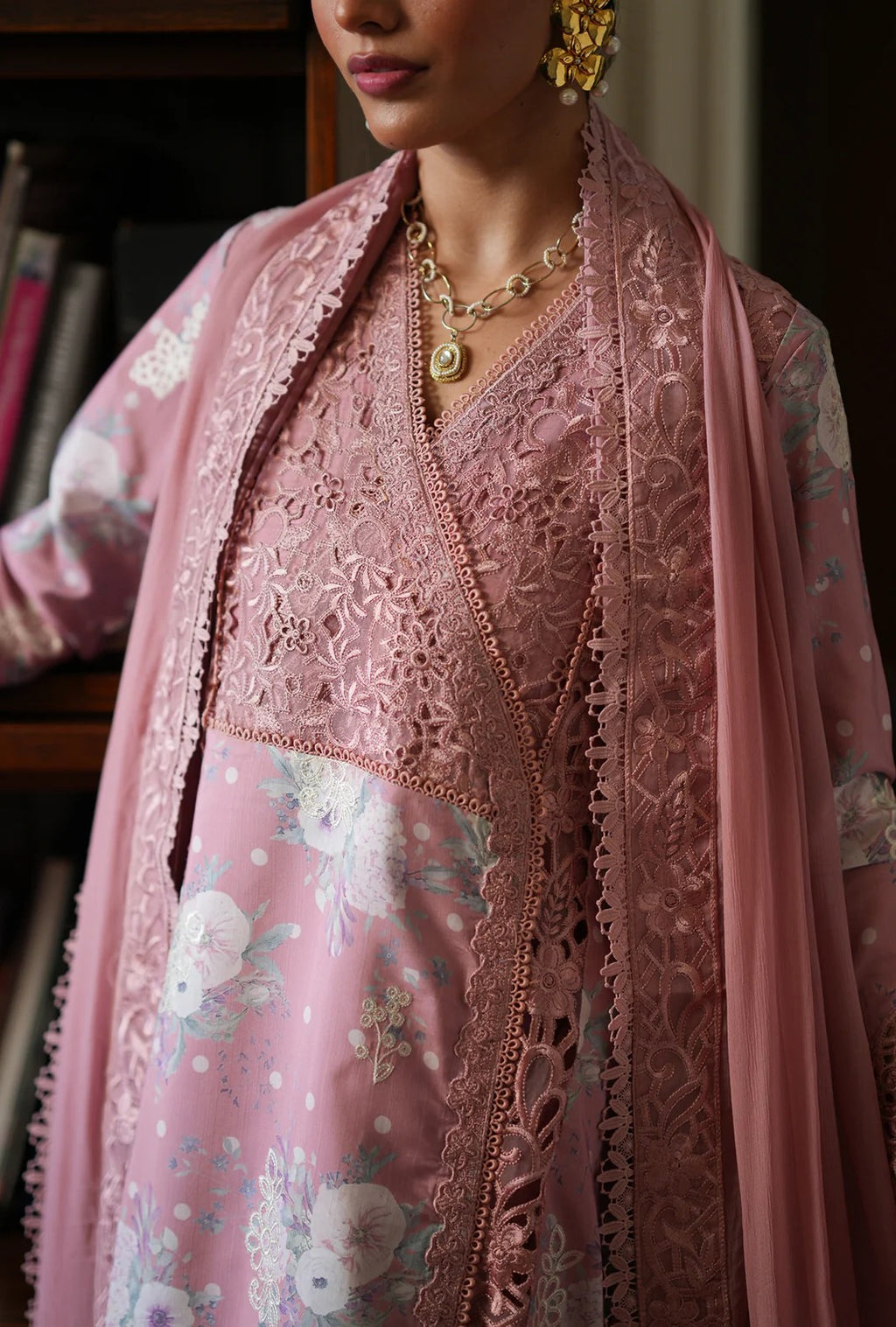 A close-up of a woman wearing a pink traditional outfit with floral embroidery and lace detailing, paired with a matching sheer dupatta and gold jewelry.