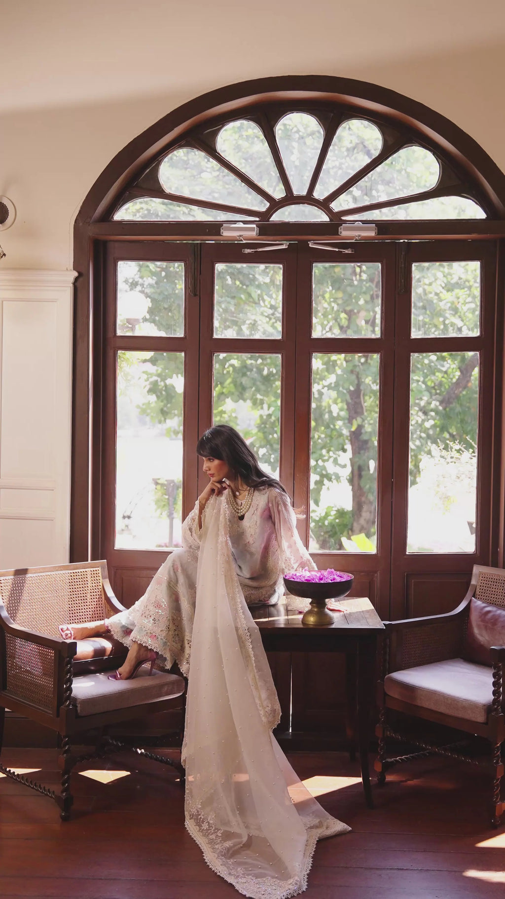 A woman in a flowing white traditional outfit sits on a wooden armchair in a sunlit room with a large arched window, dark wood furniture, and greenery visible outside.