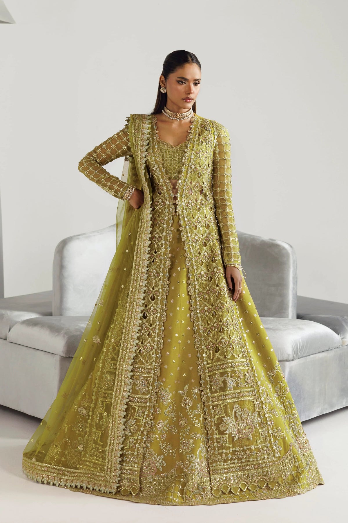 A woman models a traditional Indian anarkali gown in olive green with intricate gold embroidery and a matching embroidered dupatta, posed in front of a light gray sofa.