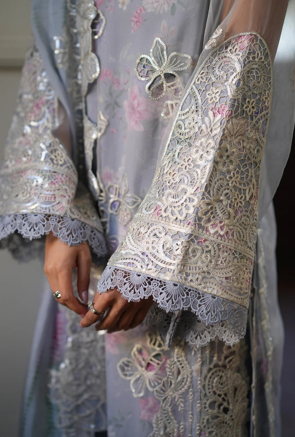A close-up of a person wearing a light lavender-gray traditional embroidered robe with ornate silver lace sleeves, floral embroidery, and delicate metallic embellishments. The person's hands are visible, adorned with rings.
