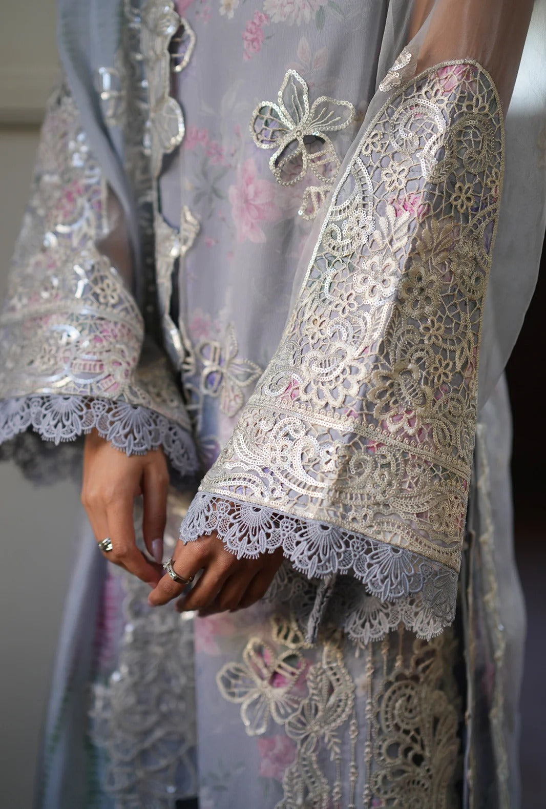 A close-up of a person wearing a light lavender-gray traditional embroidered robe with ornate silver lace sleeves, floral embroidery, and delicate metallic embellishments. The person's hands are visible, adorned with rings.