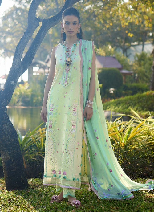 A woman stands outdoors in a garden wearing a light pastel green traditional outfit with floral embroidery and a matching draped scarf.