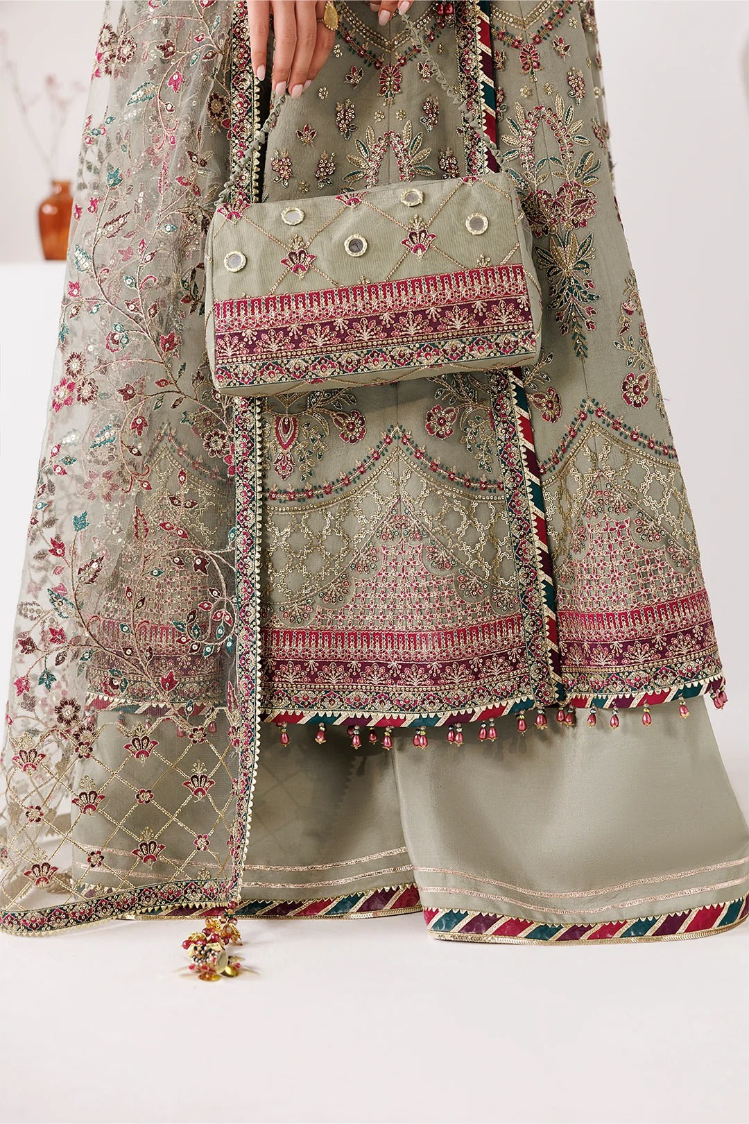 A close-up of a traditional South Asian outfit in sage green with intricate multicolor embroidery, featuring a matching embroidered clutch bag.
