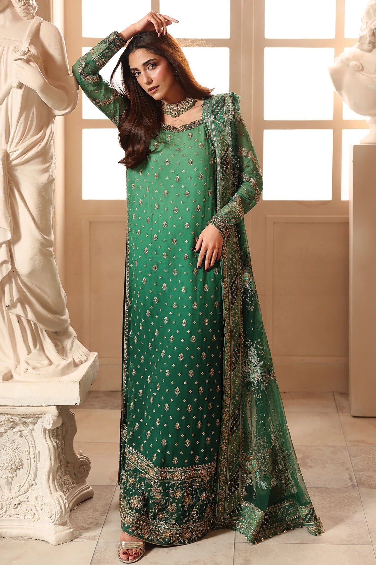 A woman models a green traditional long dress with matching embroidered dupatta in an indoor setting with classical statues.