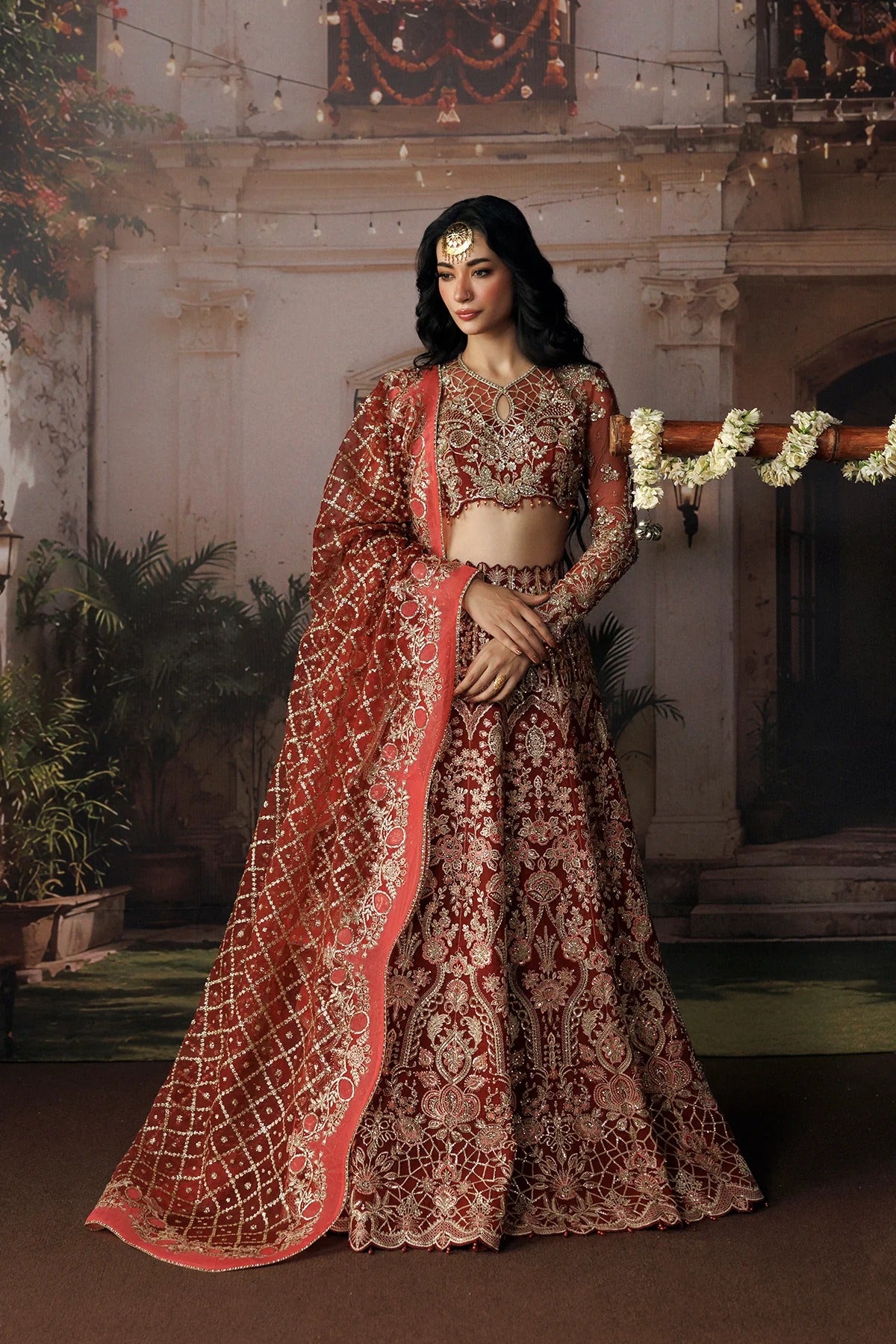 A woman models an ornate red and gold embroidered traditional Indian outfit with a matching dupatta in an indoor courtyard setting.