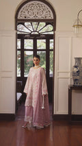 A woman models a traditional light pink embroidered kurta set with matching trousers and a dupatta in an elegant indoor setting with a wooden arched doorway.
