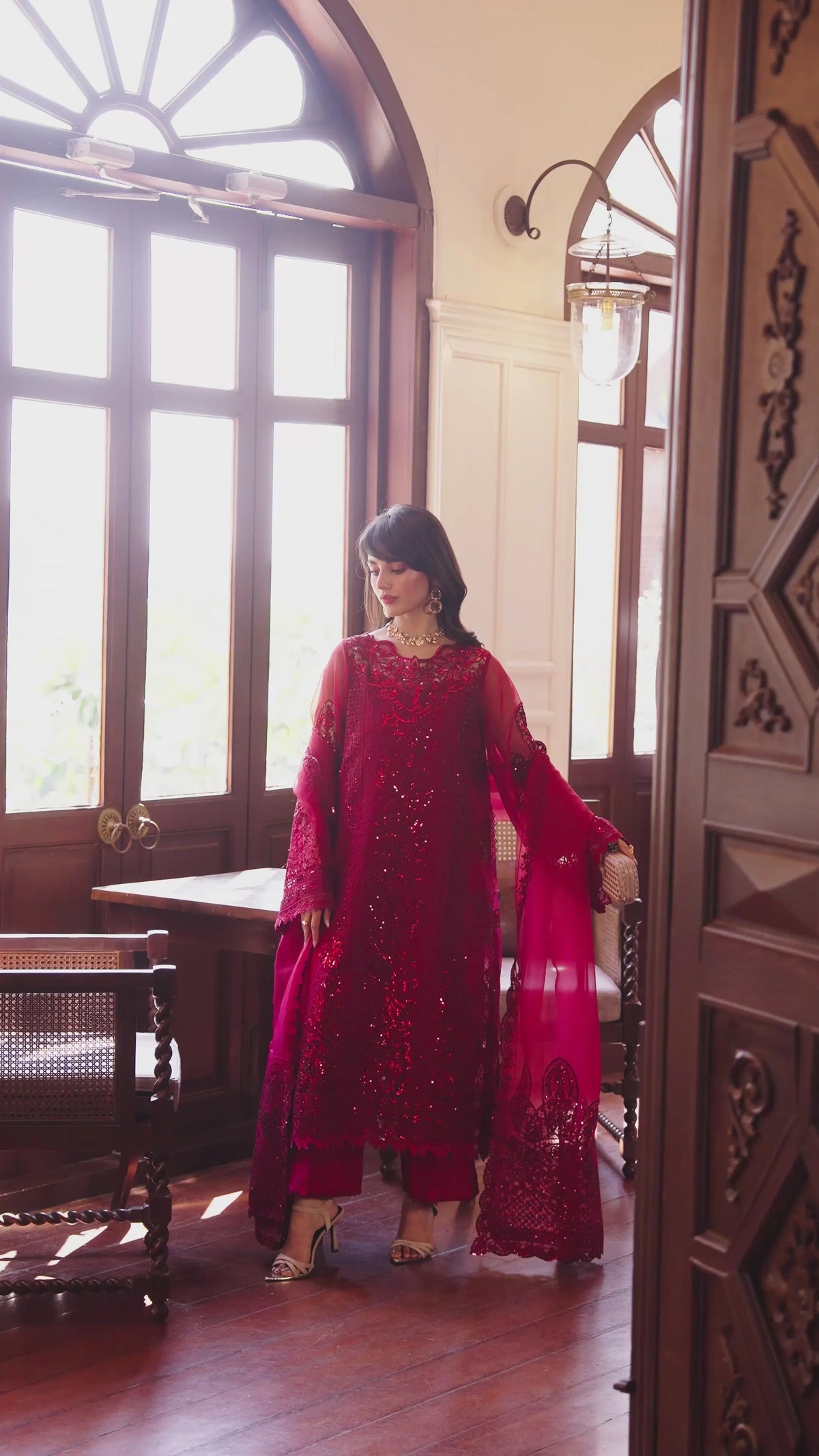 A woman wearing a deep red embroidered traditional outfit with a matching dupatta stands in a sunlit room with large arched windows and wooden furniture.
