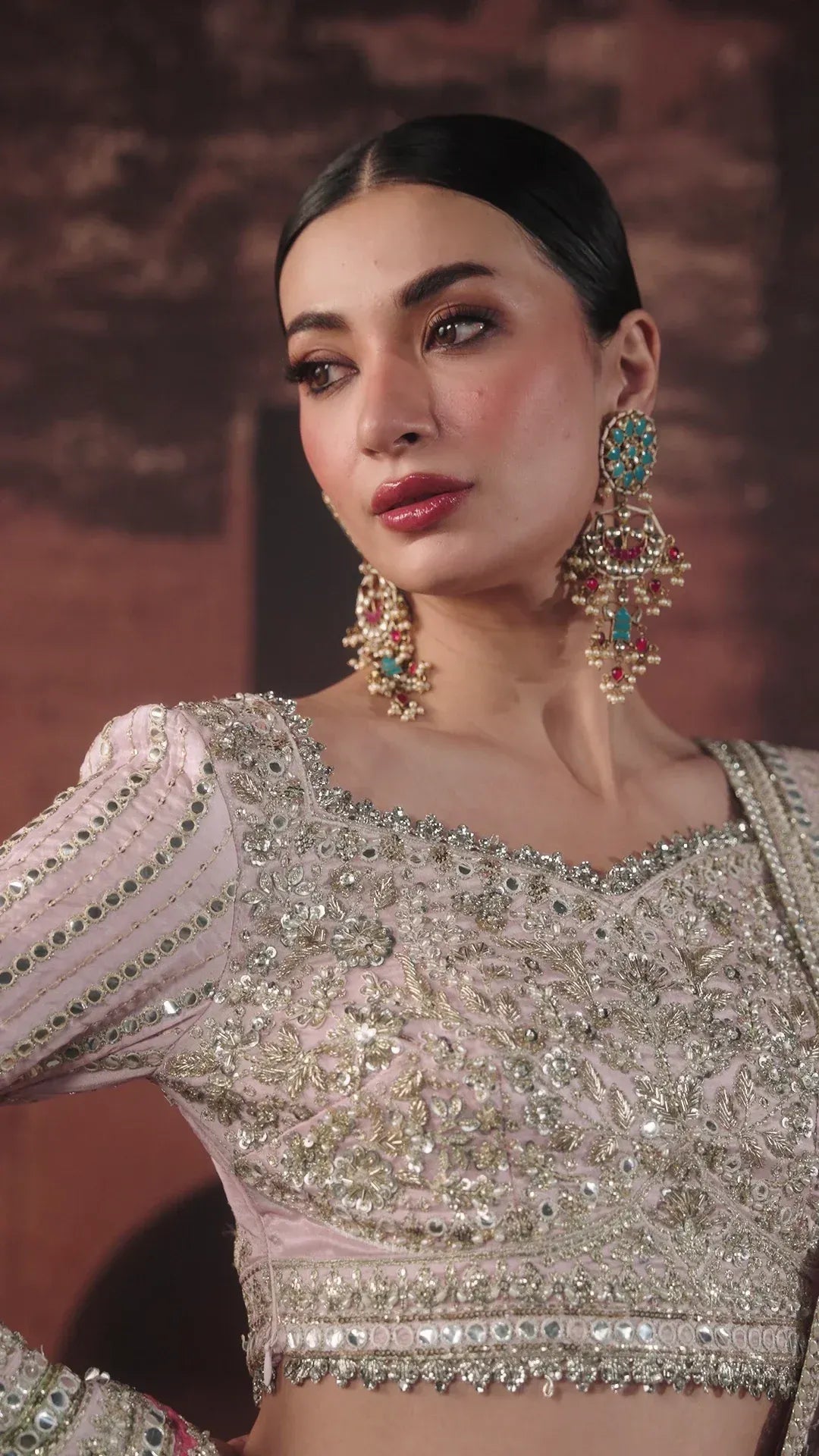 A close-up of a woman wearing an ornate light pink embellished blouse with intricate silver embroidery and beadwork, paired with matching jewelry against a warm brown background.