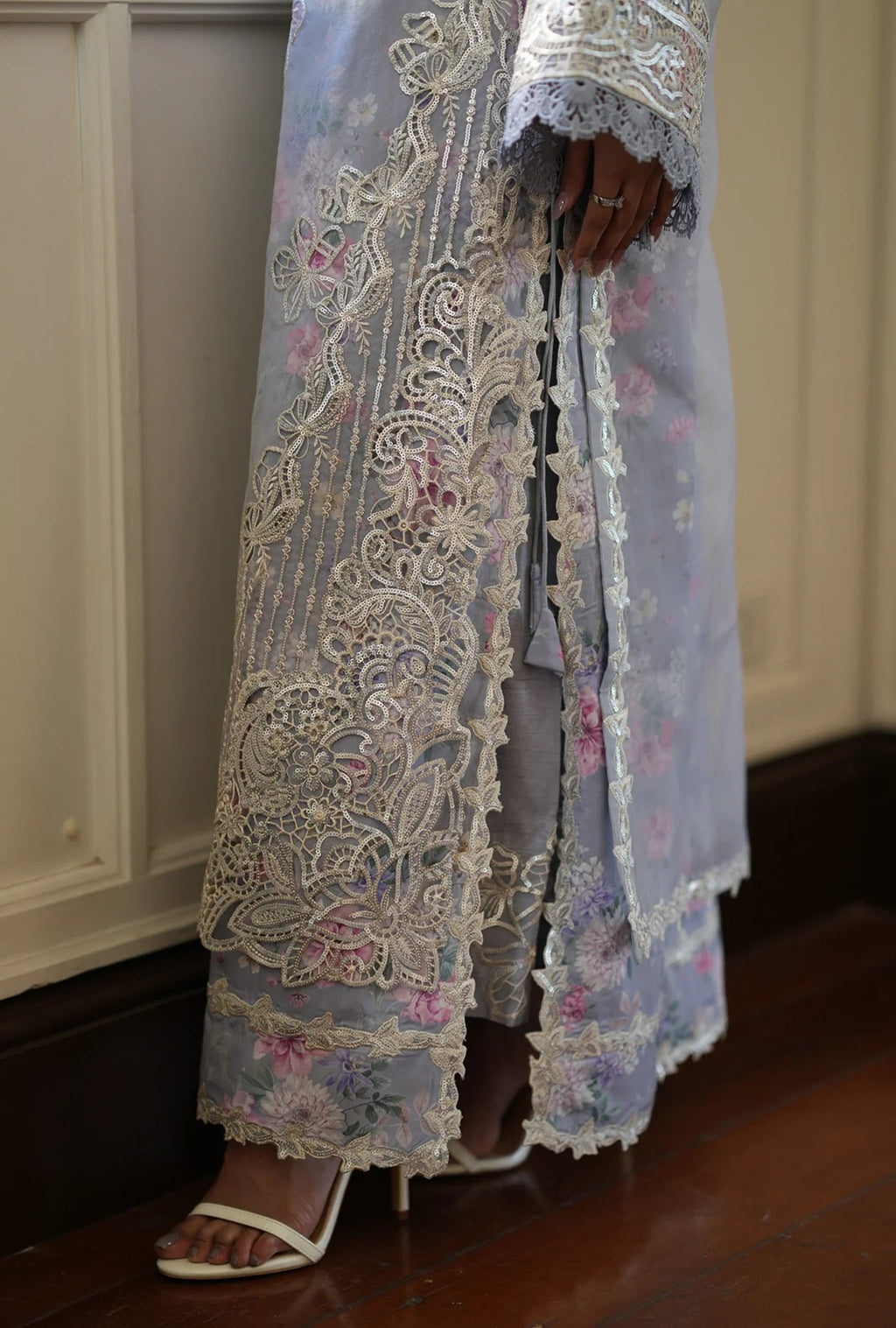 A close-up of a person wearing a sheer light blue embroidered robe with intricate white lace and floral detailing, paired with white heeled sandals, standing on a dark wood floor.