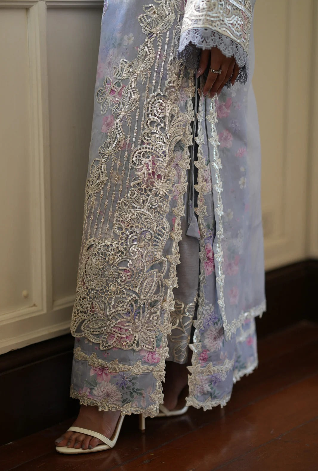 A close-up of a person wearing a sheer light blue embroidered robe with intricate white lace and floral detailing, paired with white heeled sandals, standing on a dark wood floor.