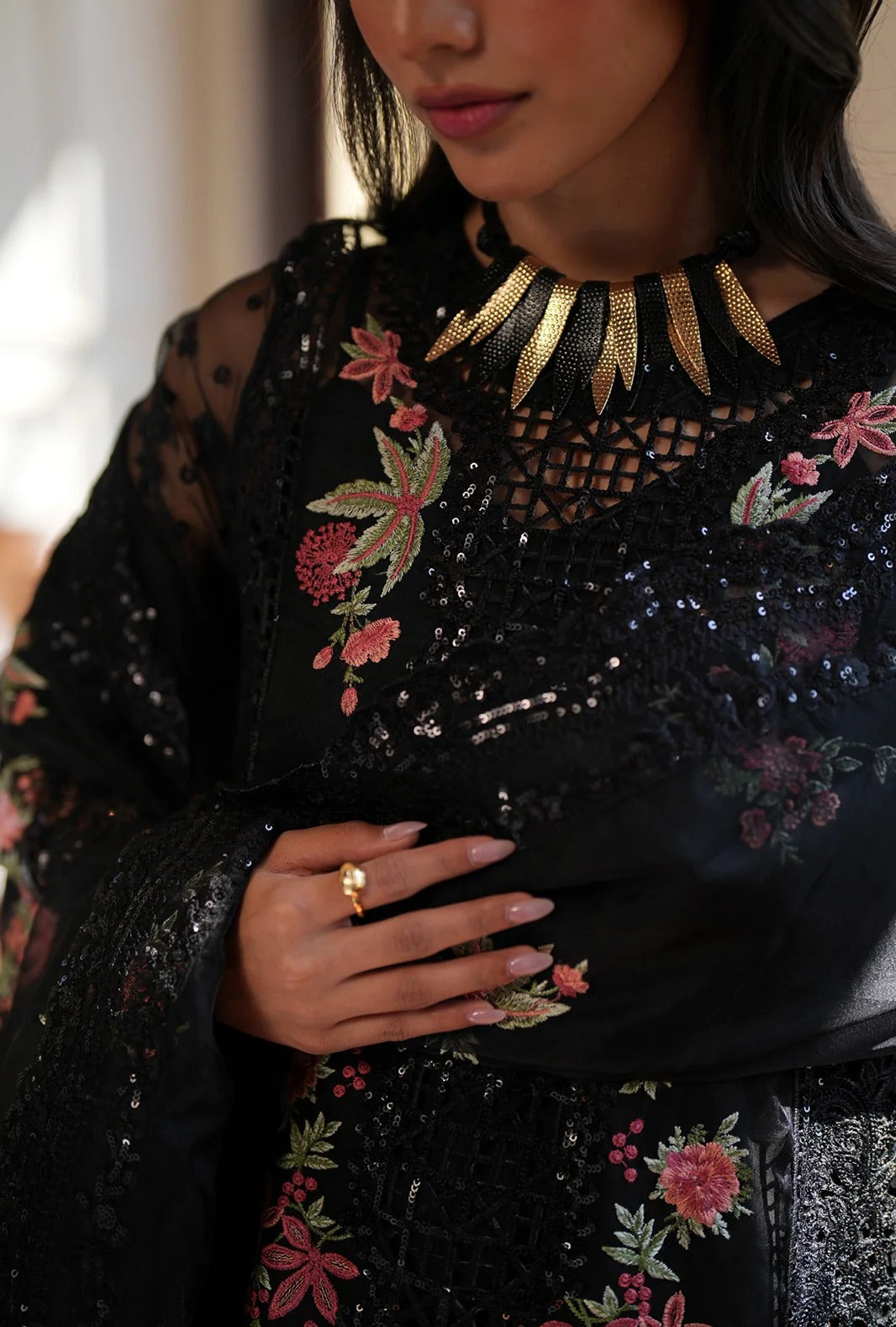 A close-up of a woman wearing a black embroidered traditional outfit with floral patterns, a gold necklace, and a gold ring, set against a softly blurred indoor background.