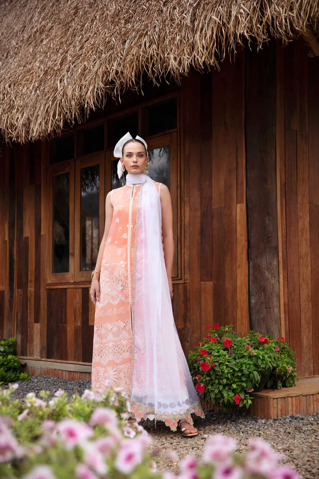 A woman stands outdoors in front of a wooden house with a thatched roof, wearing a long peach and white embroidered traditional outfit with a matching headscarf. Pink flowers are blurred in the foreground, and potted red flowers sit near the building.