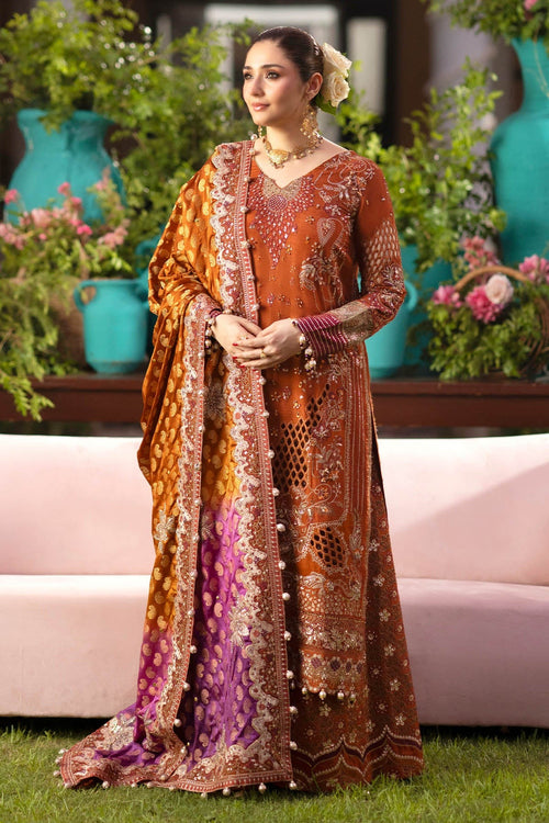 A woman models a traditional South Asian salwar kameez outfit with a matching dupatta. The outfit is primarily rust-orange with intricate gold embroidery, a purple and gold patterned border, and a purple dupatta with gold motifs.