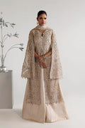 A woman modeling a long beige embroidered gown with a matching draped cape in a studio setting.