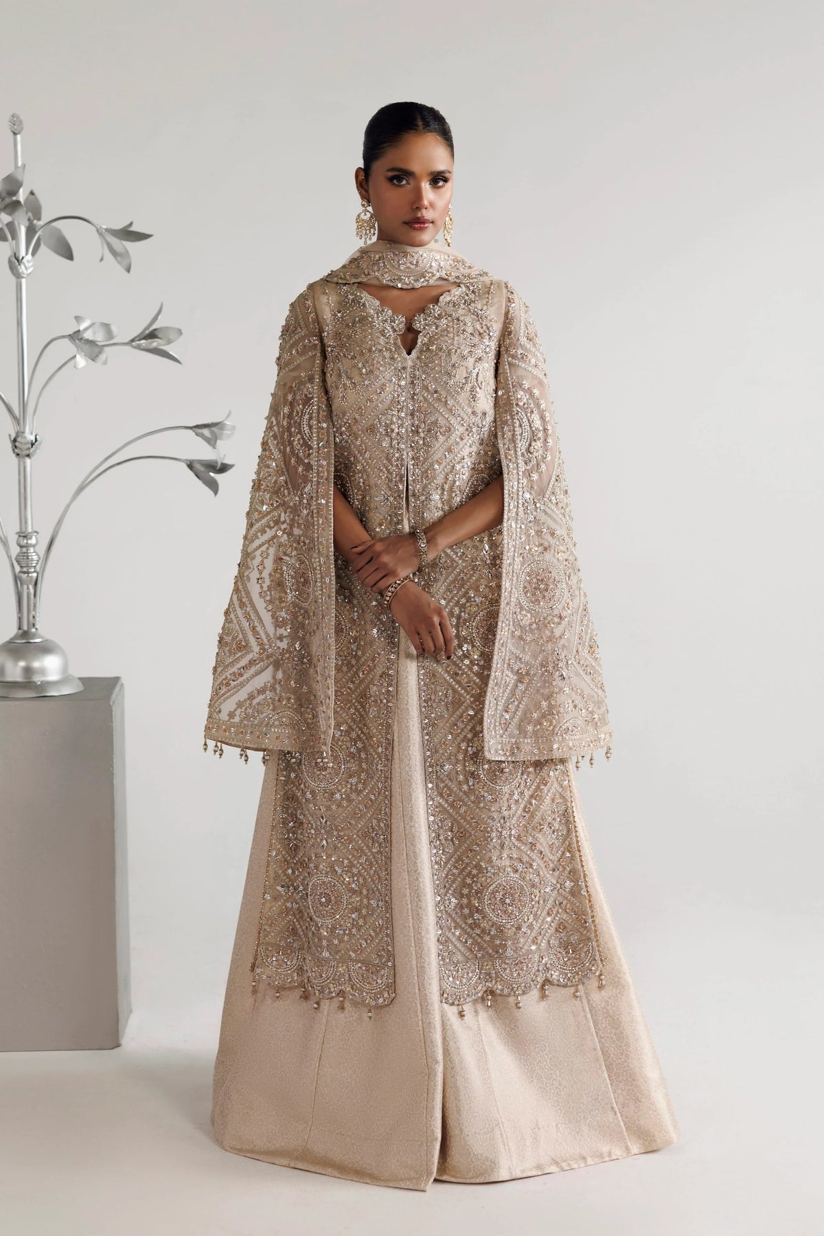 A woman modeling a long beige embroidered gown with a matching draped cape in a studio setting.