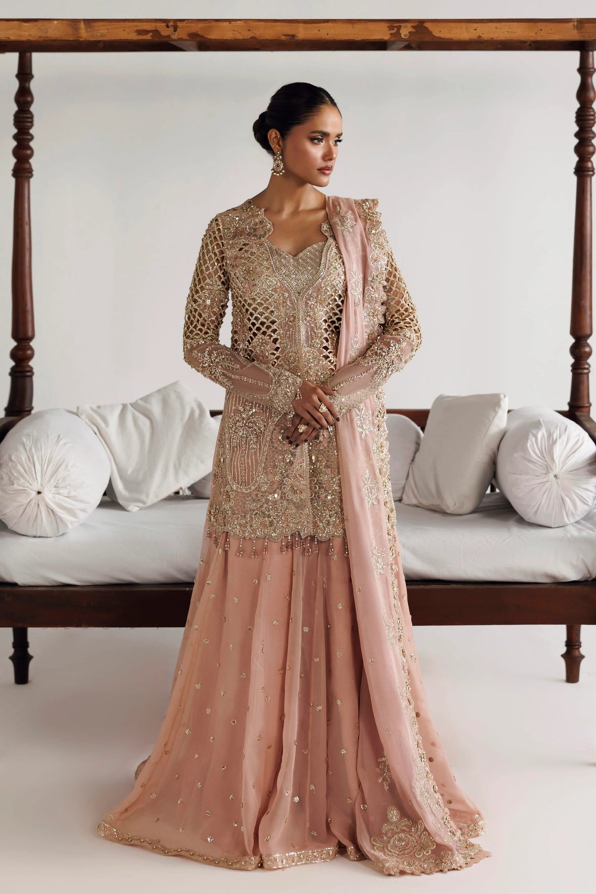 A woman models a traditional Indian salwar kameez outfit with a dusty pink skirt and a beige embroidered kameez top, draped with a matching pink dupatta, set against a white bed with wooden posts.