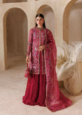 A woman models a traditional Indian outfit consisting of a heavily embroidered magenta-pink kurta, a matching pleated skirt, and a sheer dupatta with floral patterns, set against a warm beige architectural background with arches.