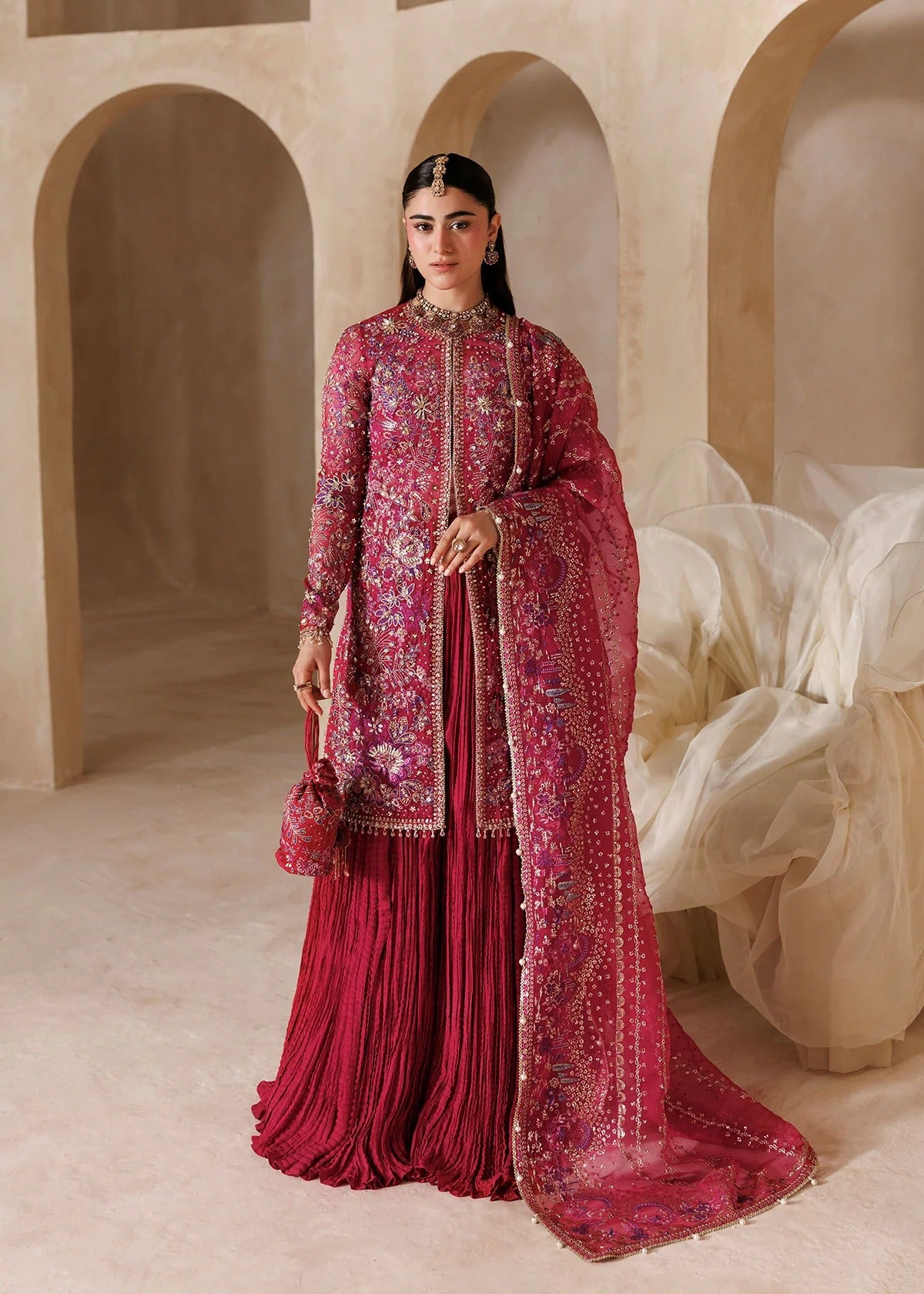A woman models a traditional Indian outfit consisting of a heavily embroidered magenta-pink kurta, a matching pleated skirt, and a sheer dupatta with floral patterns, set against a warm beige architectural background with arches.