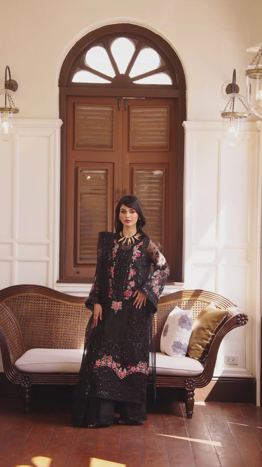A woman stands indoors in front of a large arched wooden door, wearing a long black embroidered dress with floral detailing. A woven bench with white cushions is behind her, and wall lamps are visible on both sides.