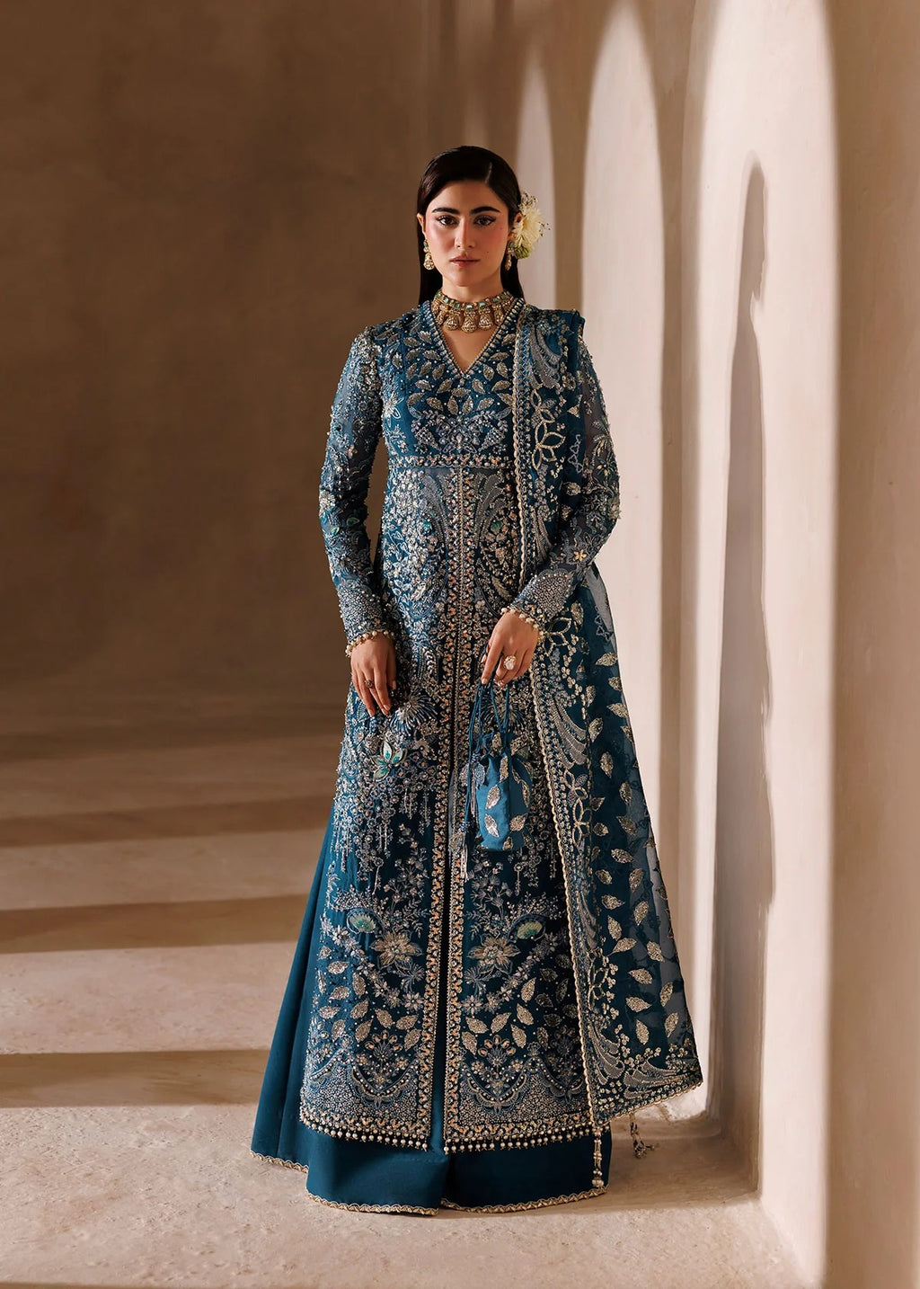 A woman stands in a softly lit architectural corridor wearing an ornate teal blue embroidered traditional gown with intricate metallic detailing and a flared skirt.