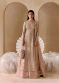 A woman stands in a studio wearing a long-sleeve, floor-length pink and beige embellished traditional outfit with a matching skirt and holding a small beige handbag. The background features warm beige arched walls and white draped fabric.