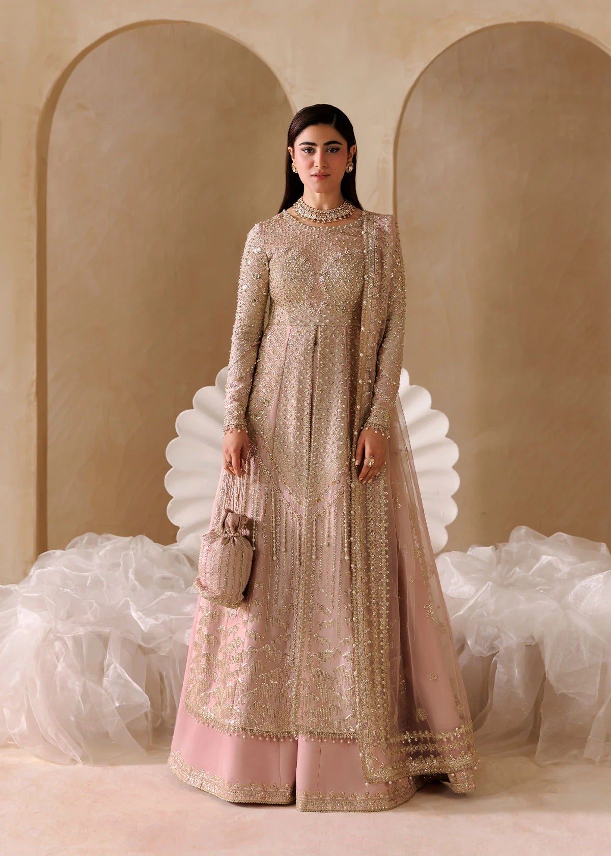 A woman stands in a studio wearing a long-sleeve, floor-length pink and beige embellished traditional outfit with a matching skirt and holding a small beige handbag. The background features warm beige arched walls and white draped fabric.