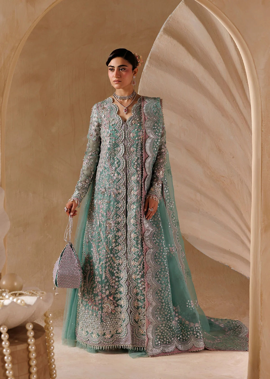 A woman models a traditional long-sleeve mint green and silver embellished gown with a matching sheer dupatta, standing in a warm beige arched interior setting.