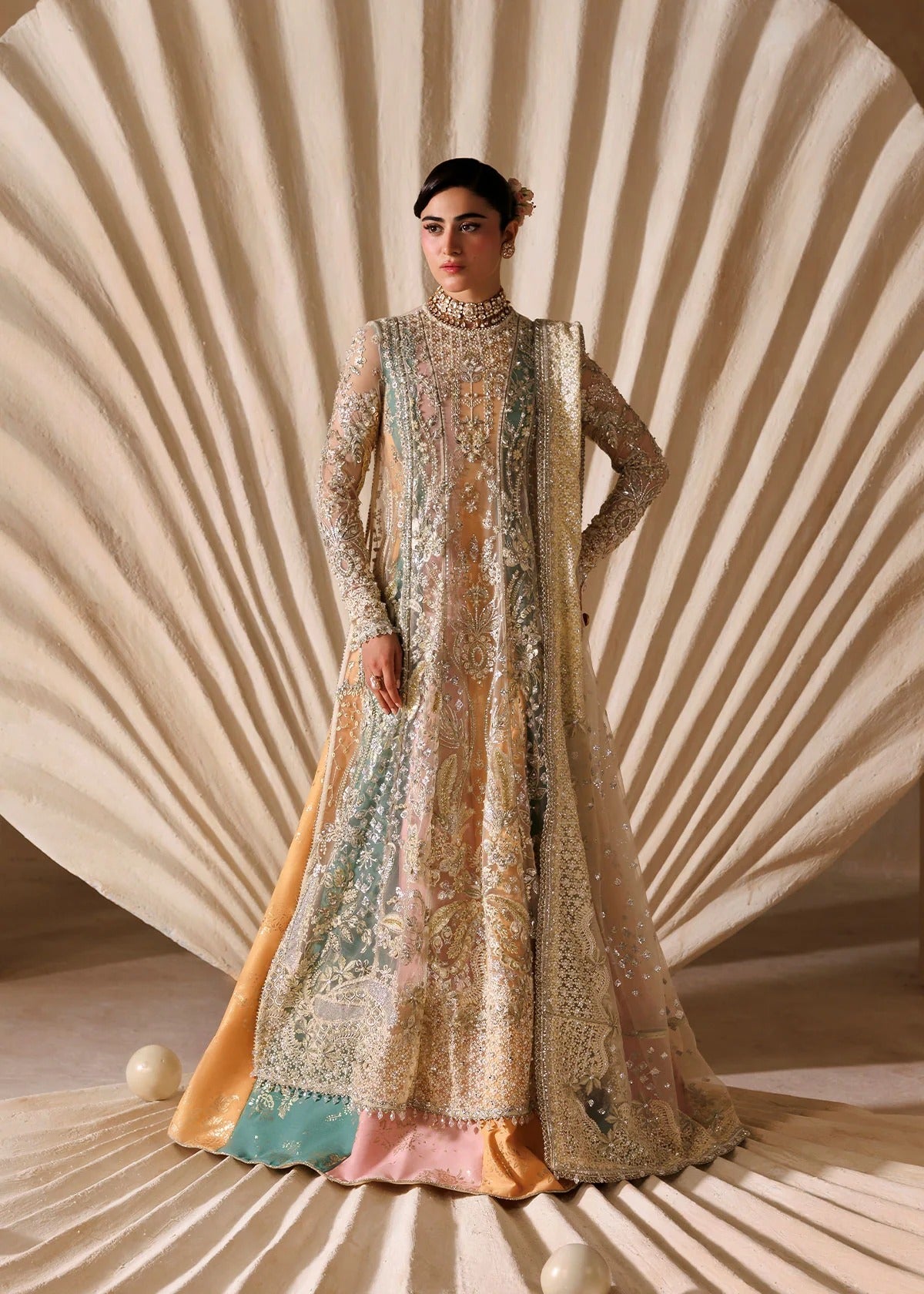 A model stands in a studio wearing an ornate, heavily embroidered traditional gown with a matching sheer draped overlay. The dress features intricate beadwork and sequins in beige, gold, light blue, and soft pink, with a solid peach-orange underlayer visible at the hem.