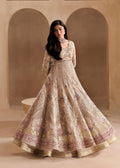A woman modeling a heavily embellished beige and gold traditional gown with intricate embroidery, standing in front of a warm beige arched backdrop.