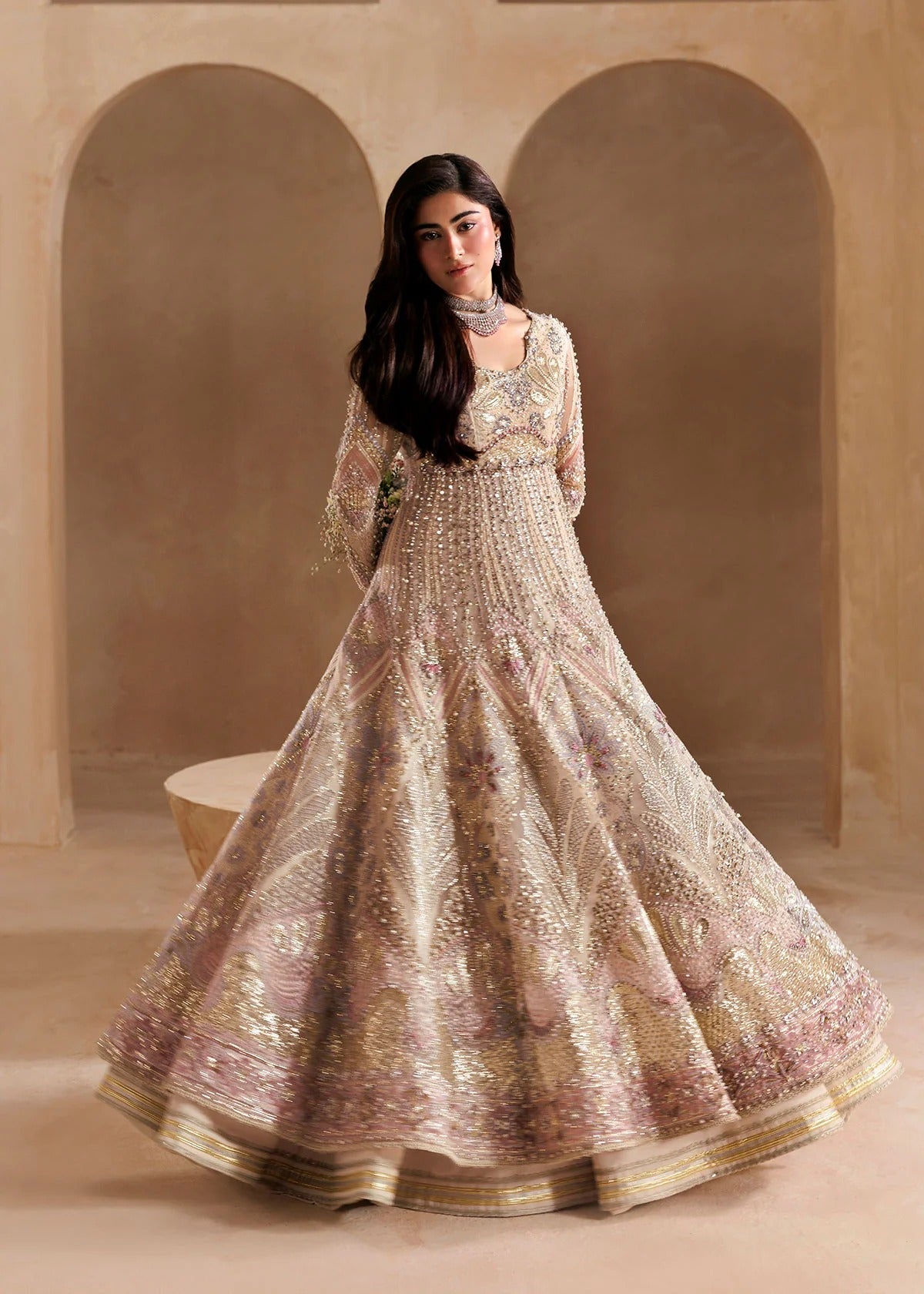 A woman modeling a heavily embellished beige and gold traditional gown with intricate embroidery, standing in front of a warm beige arched backdrop.