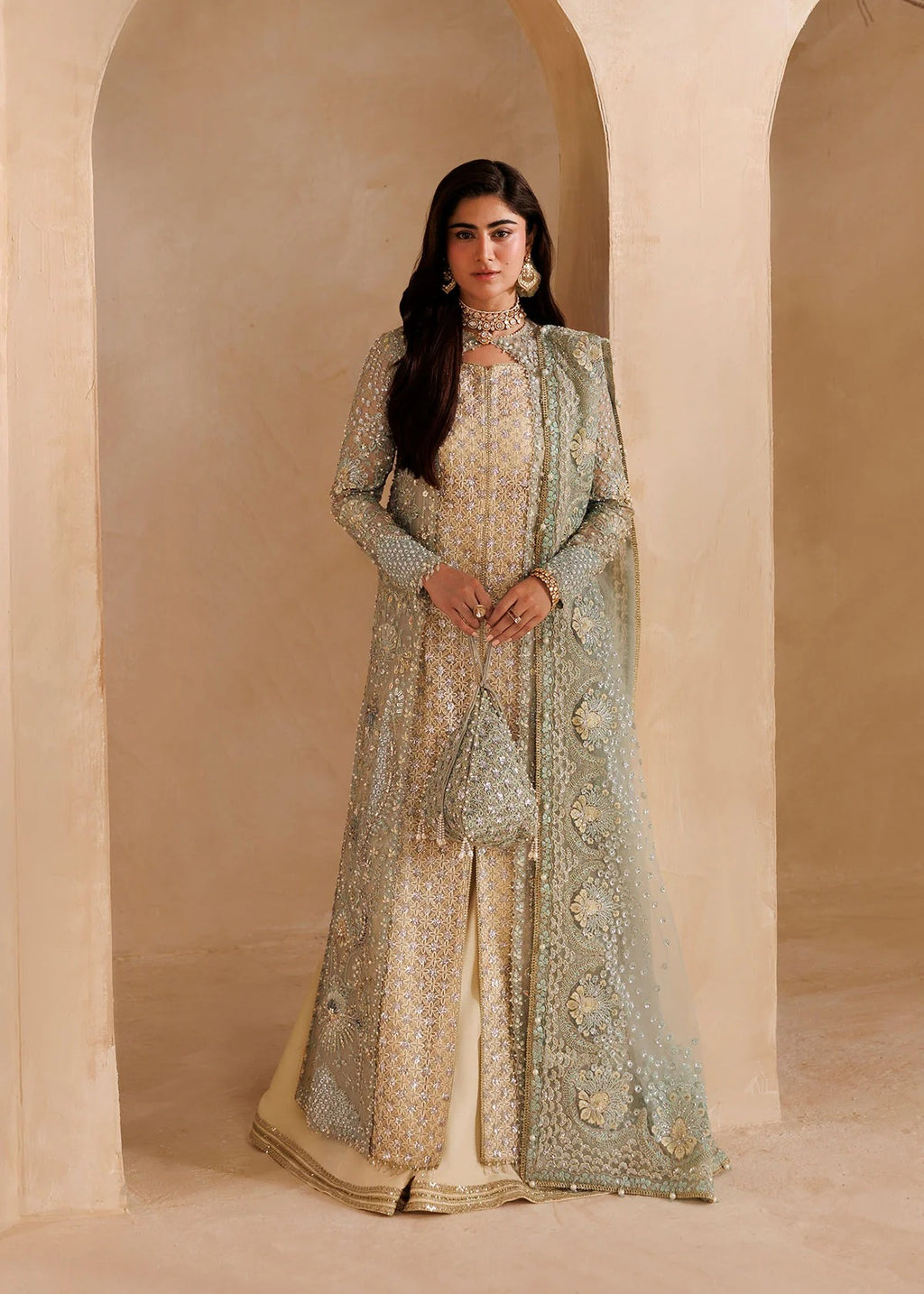 A woman stands in a warm beige arched interior wearing an ornate traditional outfit with a long cream base and heavy silver embellishment, paired with a sheer pale green draped overlay and matching clutch.