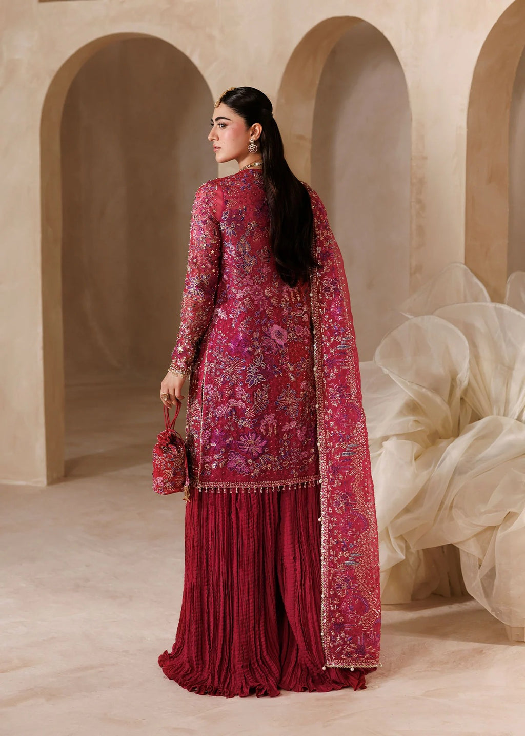 A woman wearing a traditional embroidered salwar kameez set in deep magenta and burgundy, standing in a warm beige architectural setting with arches.