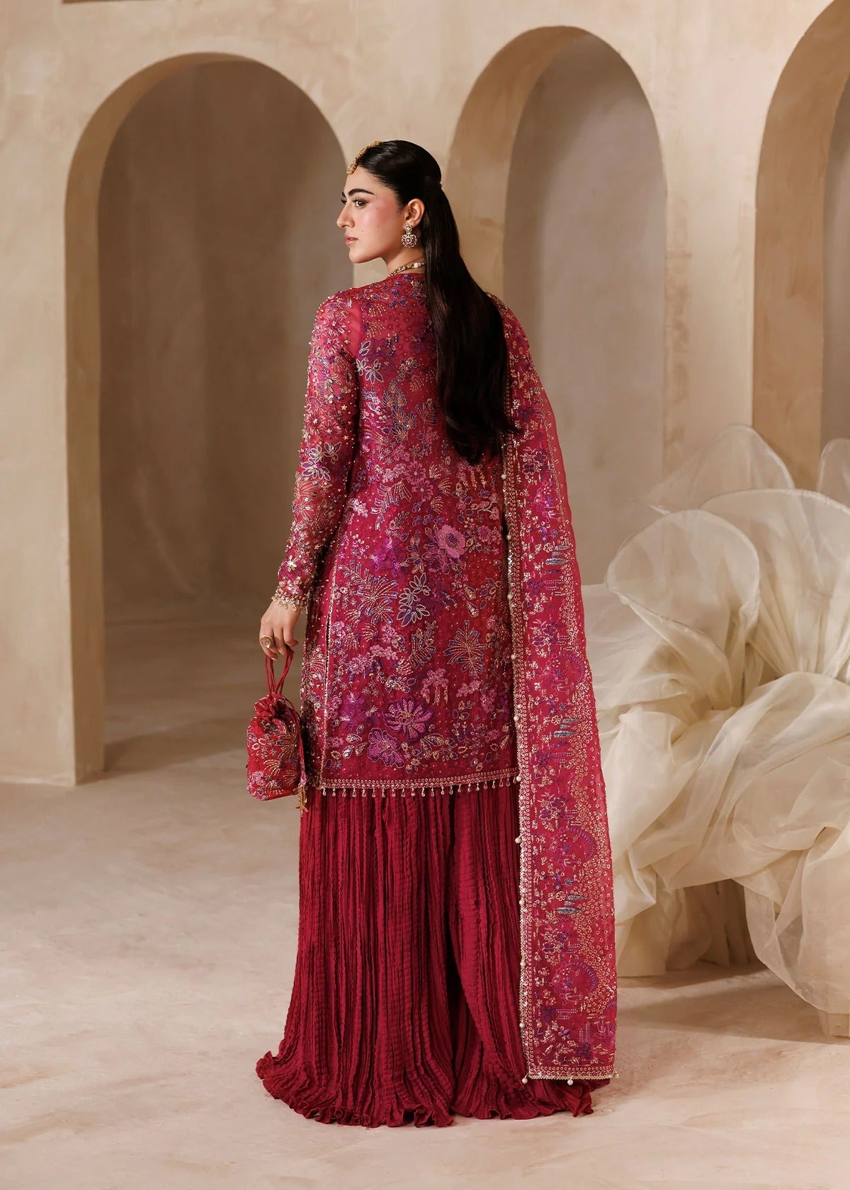 A woman wearing a traditional embroidered salwar kameez set in deep magenta and burgundy, standing in a warm beige architectural setting with arches.