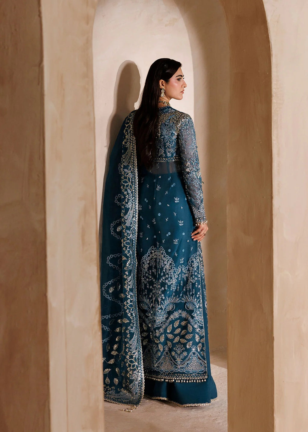 A woman wearing a dark teal blue traditional embroidered dress with a matching dupatta, standing in a warm beige stone archway.