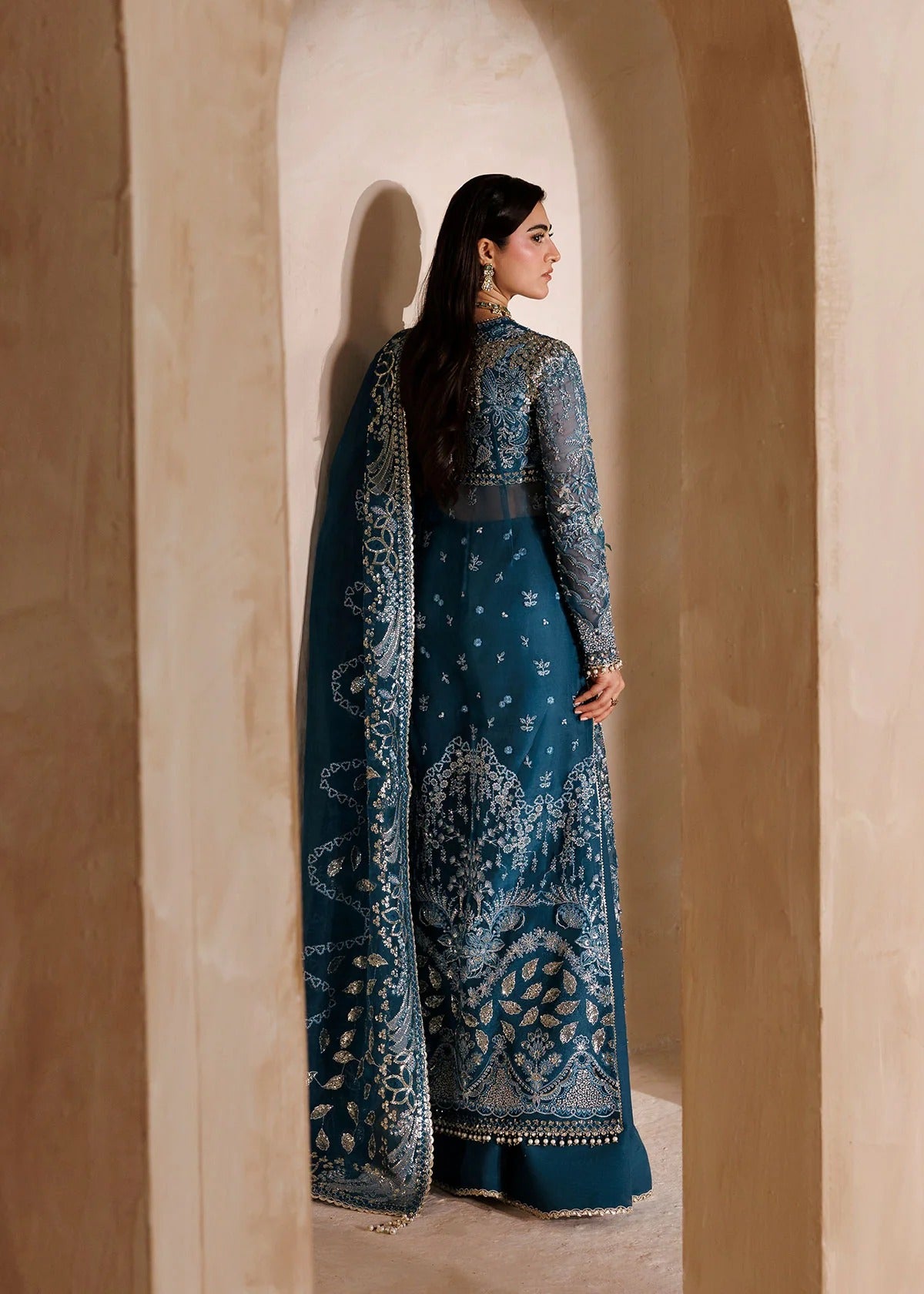 A woman wearing a dark teal blue traditional embroidered dress with a matching dupatta, standing in a warm beige stone archway.