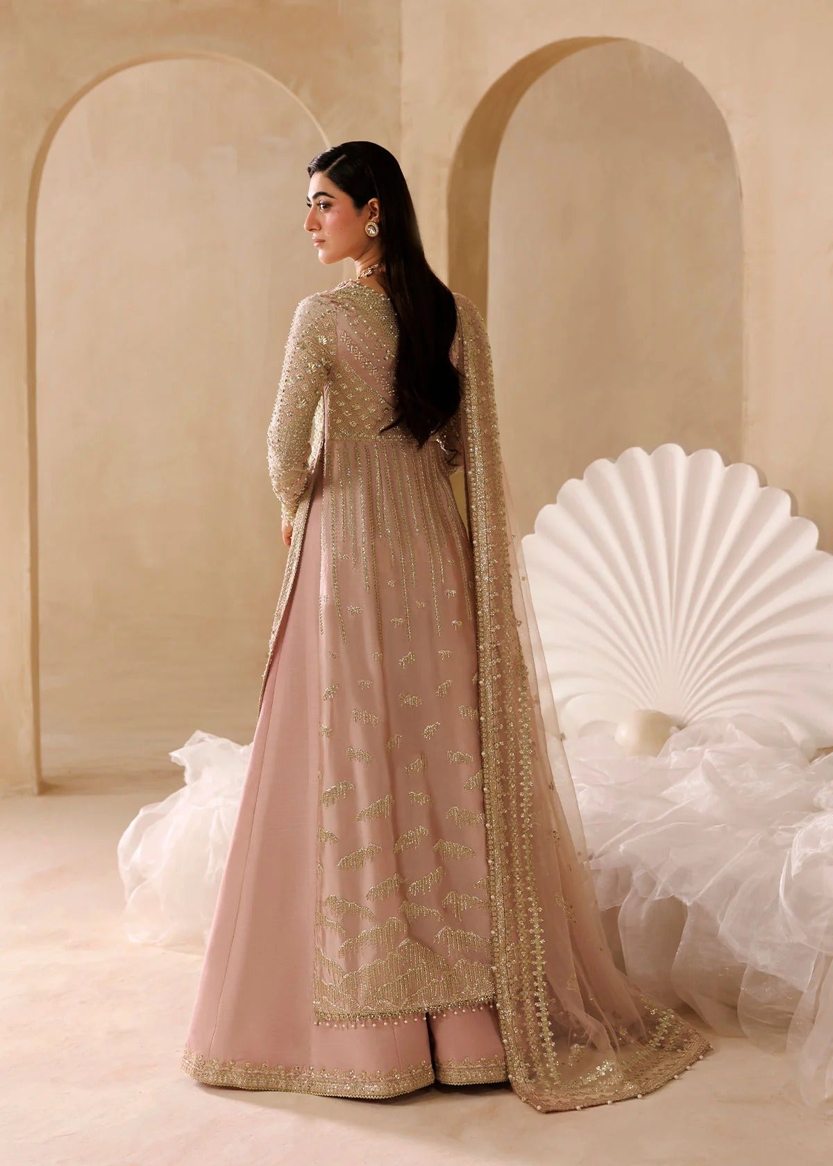 A woman wearing a traditional Indian anarkali gown in dusty pink with intricate gold embroidery, standing in a warm beige room with arched walls and a white decorative chair.