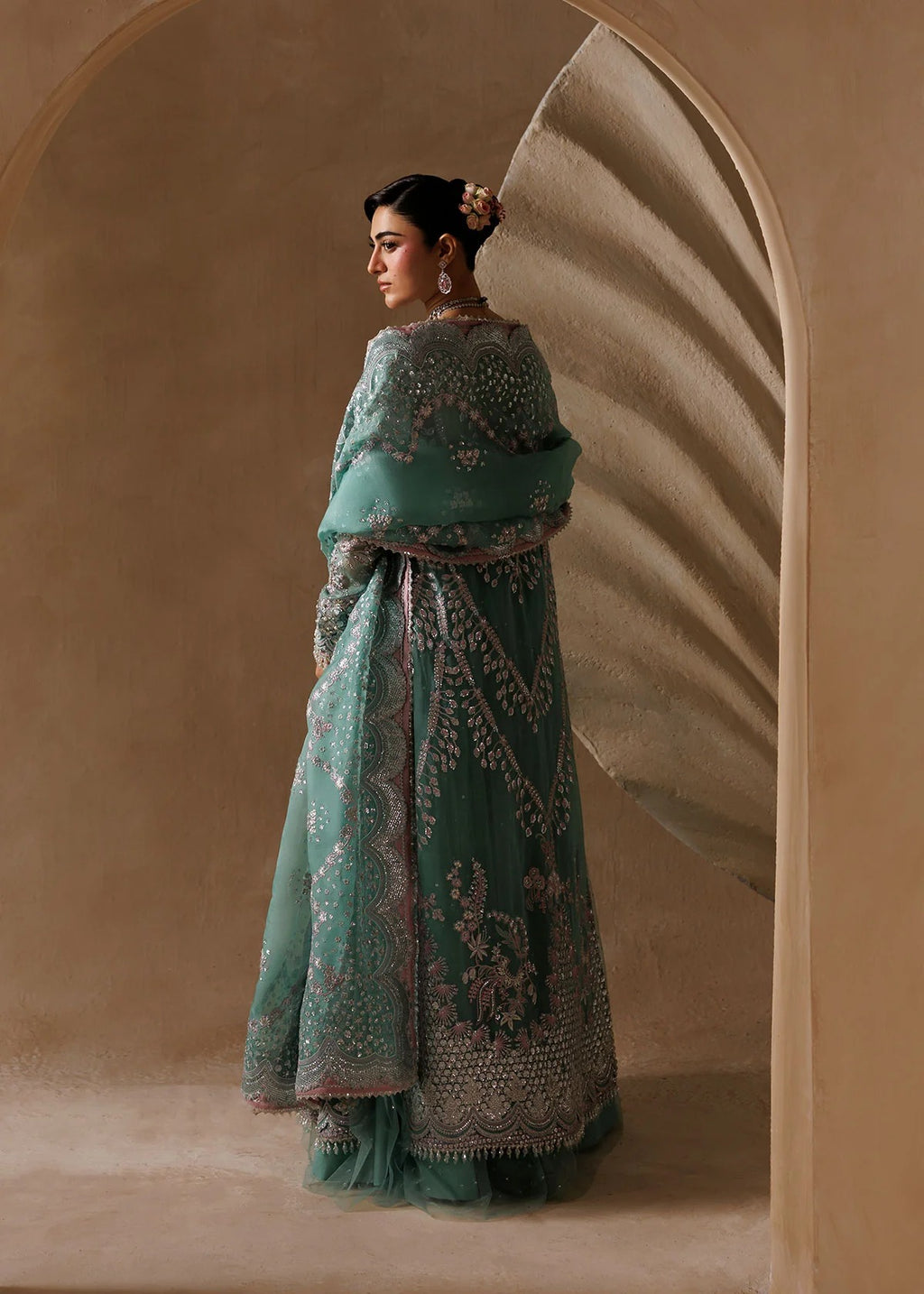 A woman wearing a traditional teal green embroidered dress with a matching dupatta, standing in a warm beige arched corridor.