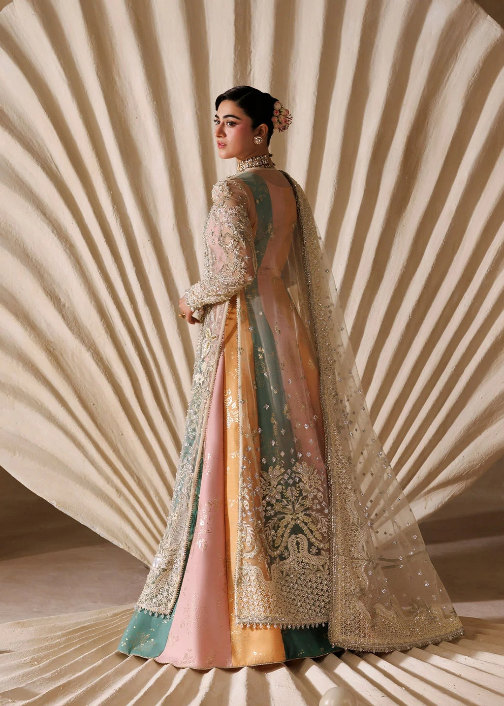 A woman models a traditional South Asian outfit with a heavily embroidered cream dupatta, a peach and sage green embroidered kurta, and a pink skirt, posed against a beige fan-like backdrop.