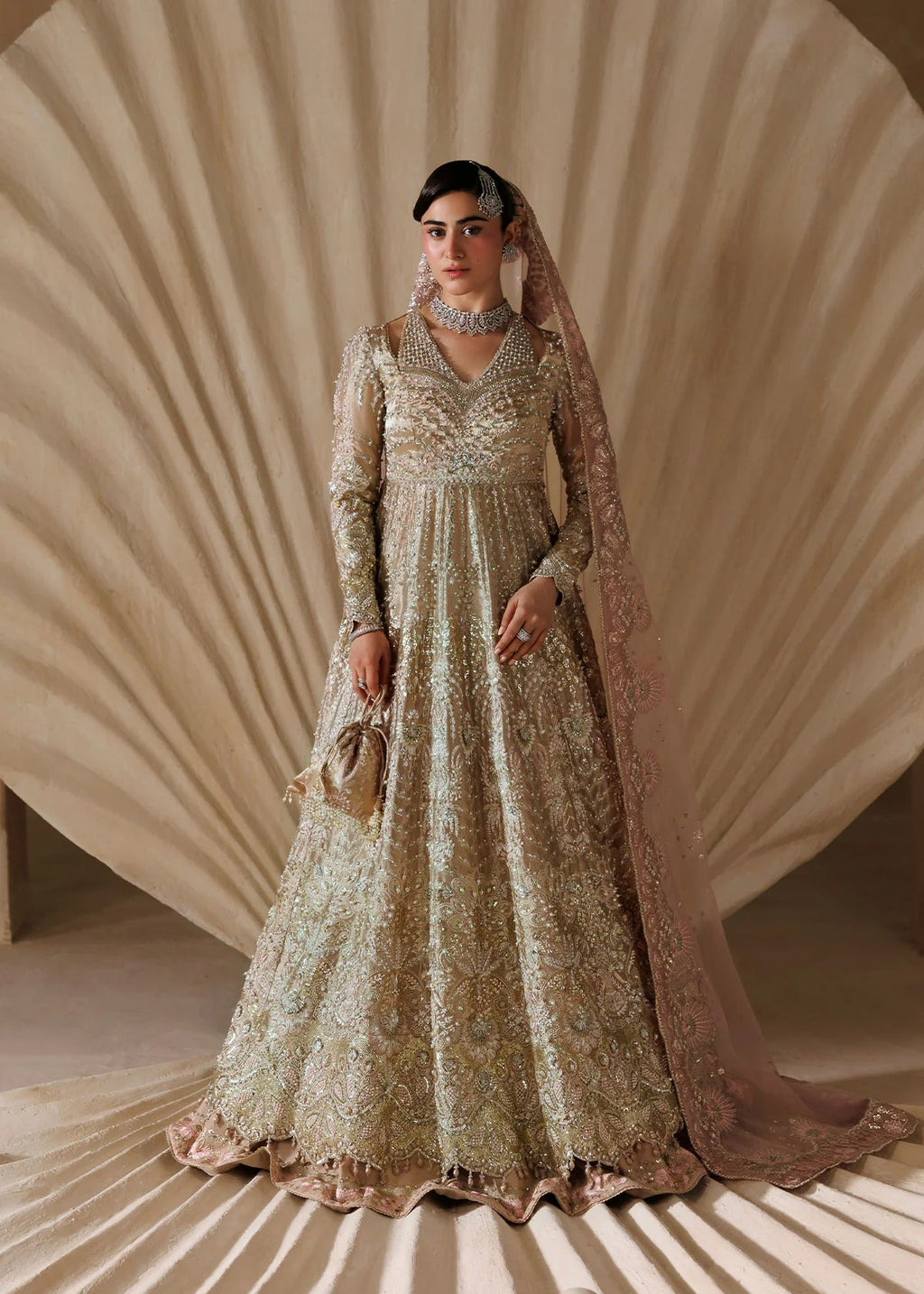 A woman models an ornate beige and gold heavily embroidered traditional gown with a matching sheer dupatta, set against a draped beige studio backdrop.