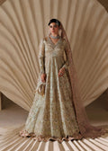 A woman models an ornate beige and gold heavily embroidered traditional gown with a matching sheer dupatta, set against a draped beige studio backdrop.