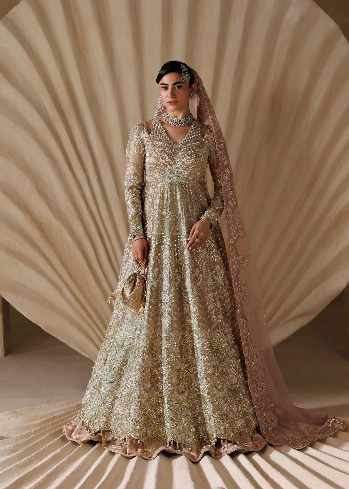 A woman models an ornate beige and gold heavily embroidered traditional gown with a matching sheer dupatta, set against a draped beige studio backdrop.